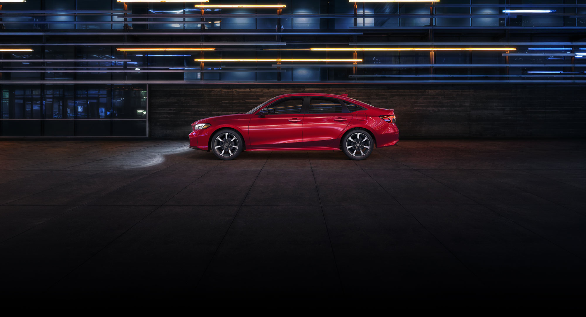 2027 Honda Civic Sedan Sport Touring Hybrid in Rallye Red profile in front of a modern building at night (Compact Sedan).
